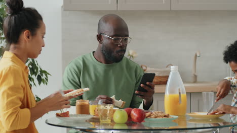 Hombre-Afroamericano-Que-Usa-Un-Teléfono-Inteligente-En-El-Desayuno-Con-Su-Familia-En-Casa